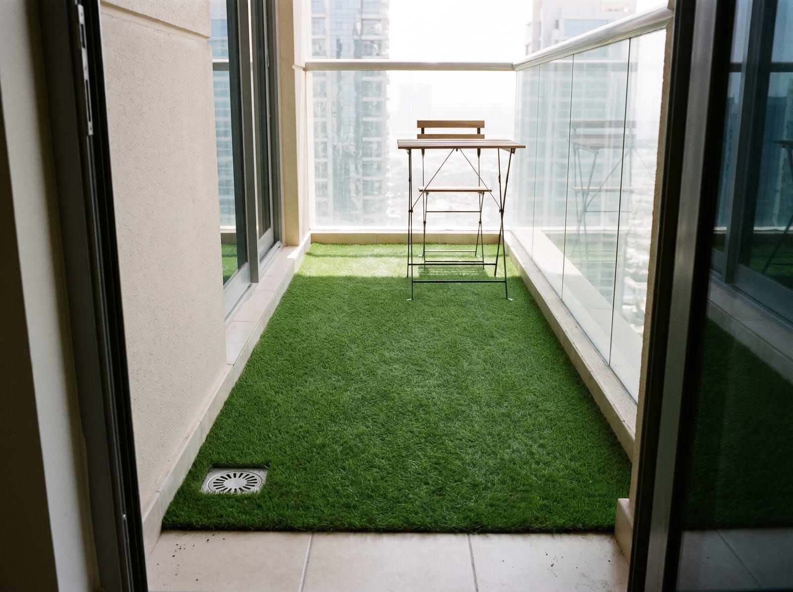 Short artificial grass laid over a high-rise balcony floor, a floor drain left clear and a bistro table at the far end