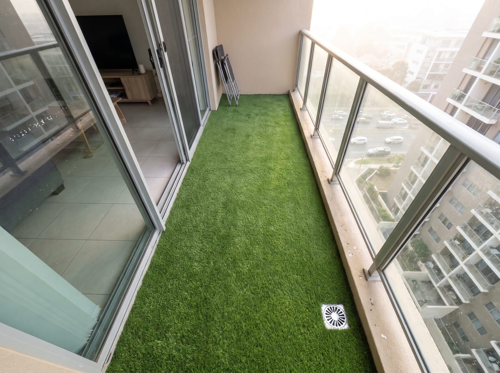 A narrow tower balcony covered in artificial grass seen from above, the sliding door to the flat open on the left and a drain at the far end