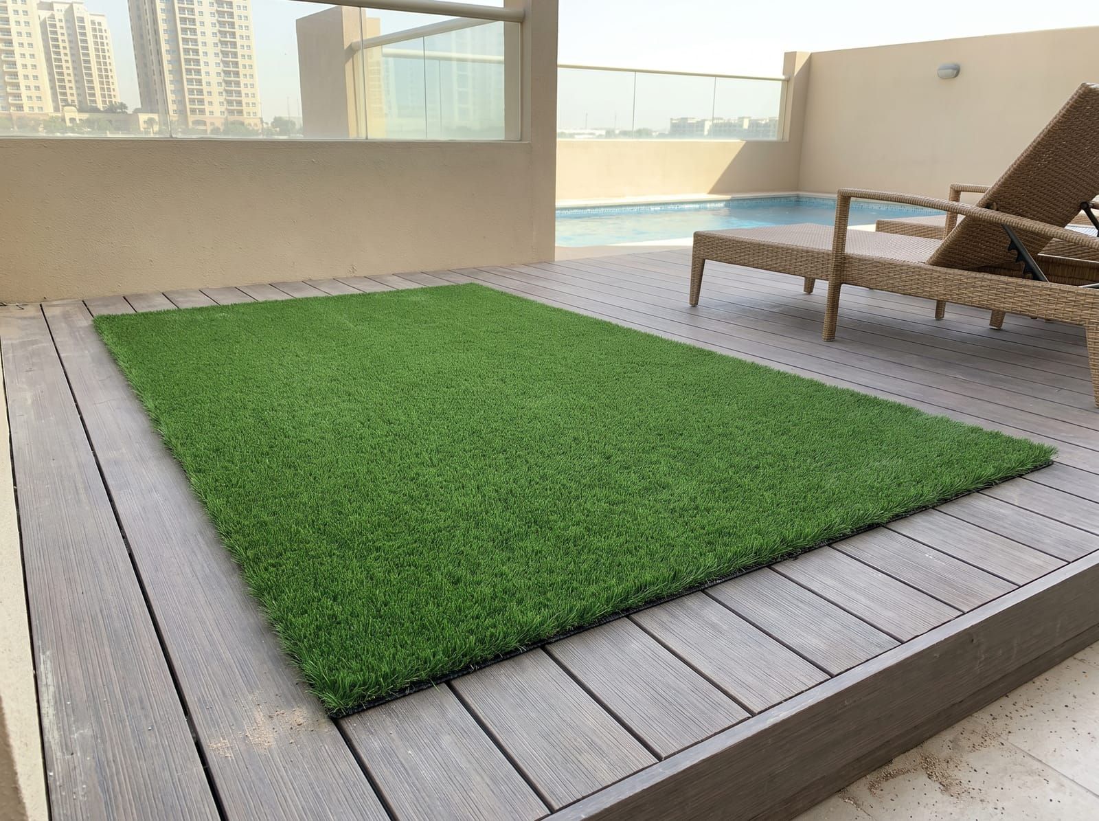 A rectangle of artificial grass laid over a composite deck with a border of bare boards, a lounger and a pool behind