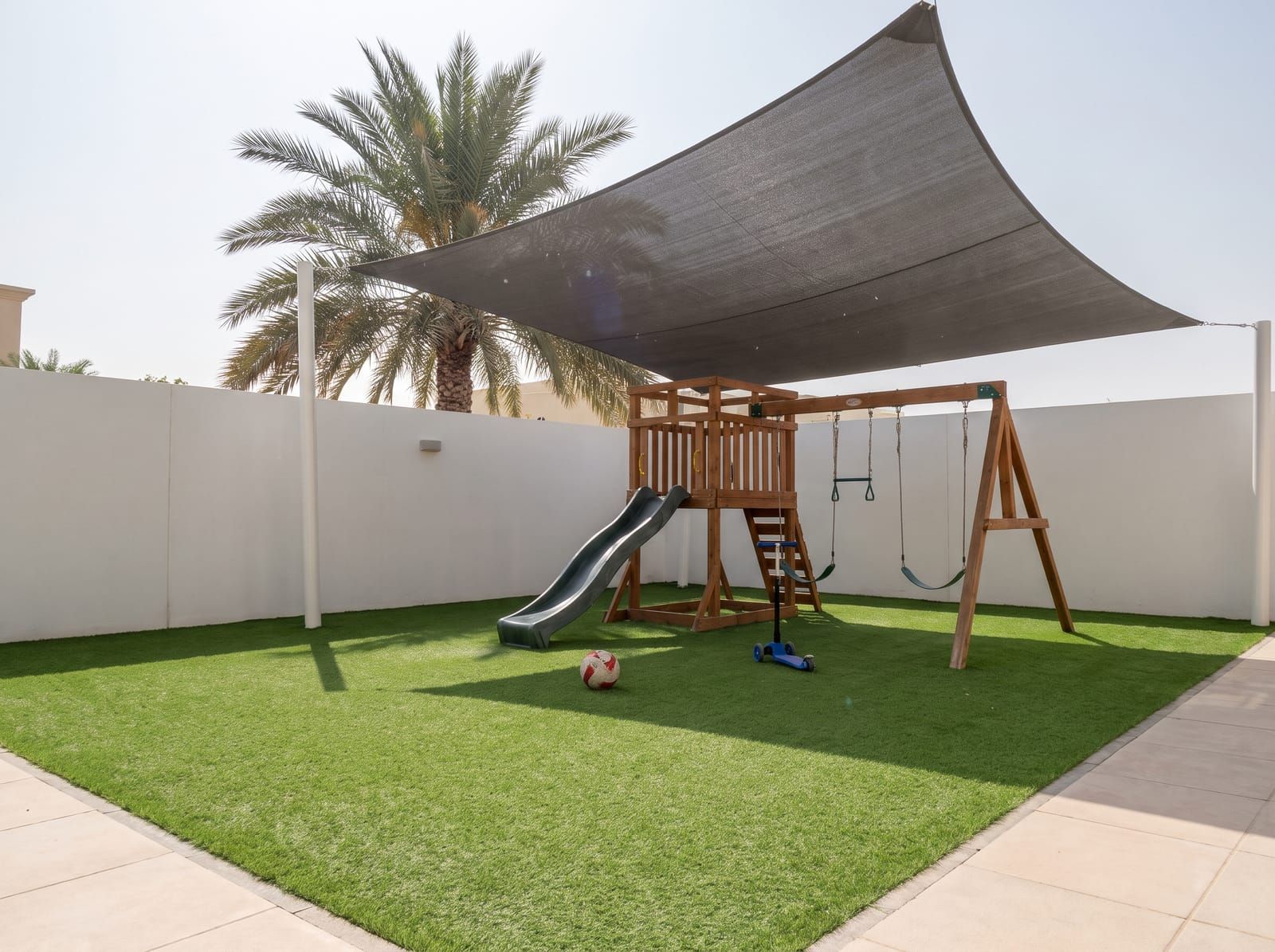 An empty play area on artificial grass, a wooden climbing frame with a slide and swings under a grey shade sail, a ball and a scooter on the lawn