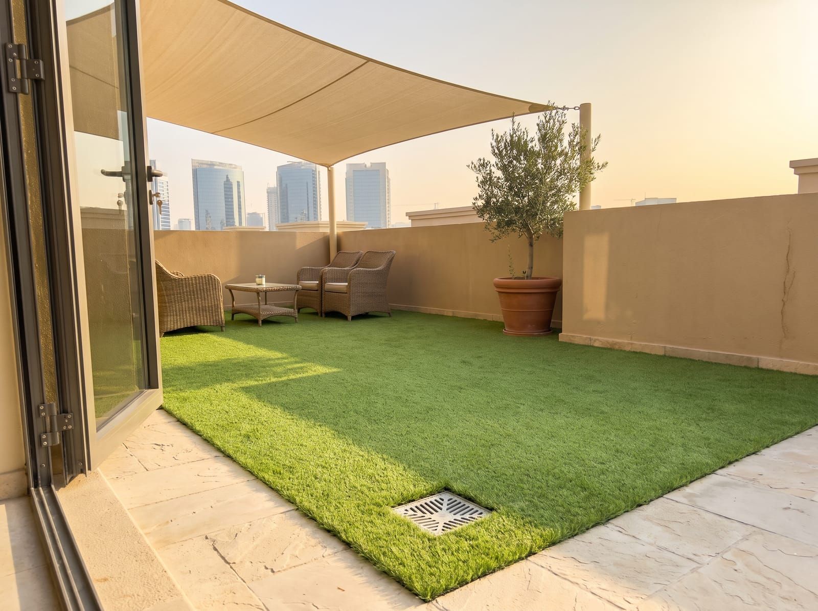 Artificial grass on a rooftop terrace under a sand-coloured shade sail, with rattan seating, an olive tree and a floor drain left clear