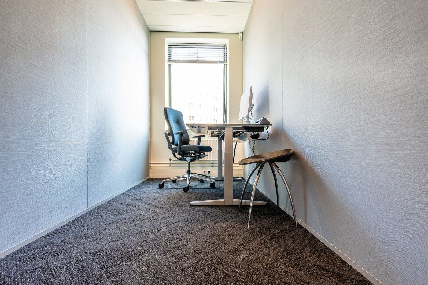 A small office with grey carpet tiles laid across the floor under a desk